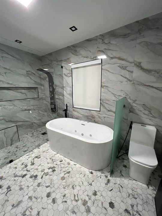 Soaking tub with integrated jets, floor-to-ceiling stone-patterned wall tile, glass-enclosed walk-in shower with built-in bench and recessed shelving, and hexagonal mosaic floor tiling
