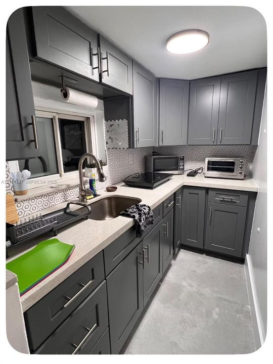 Kitchen featuring dark grey shaker-style cabinetry, light stone-composite countertops, stainless steel undermount sink with arched pull-down faucet, geometric patterned tile backsplash, and recessed ceiling lighting