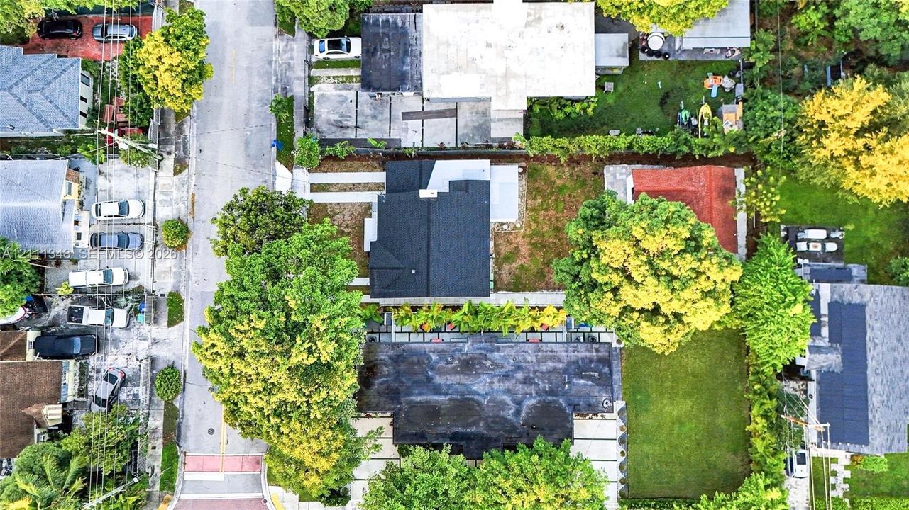 Aerial perspective featuring a multi-structure property with paved driveway access, concrete hardscaping, mature trees, and a manicured lawn