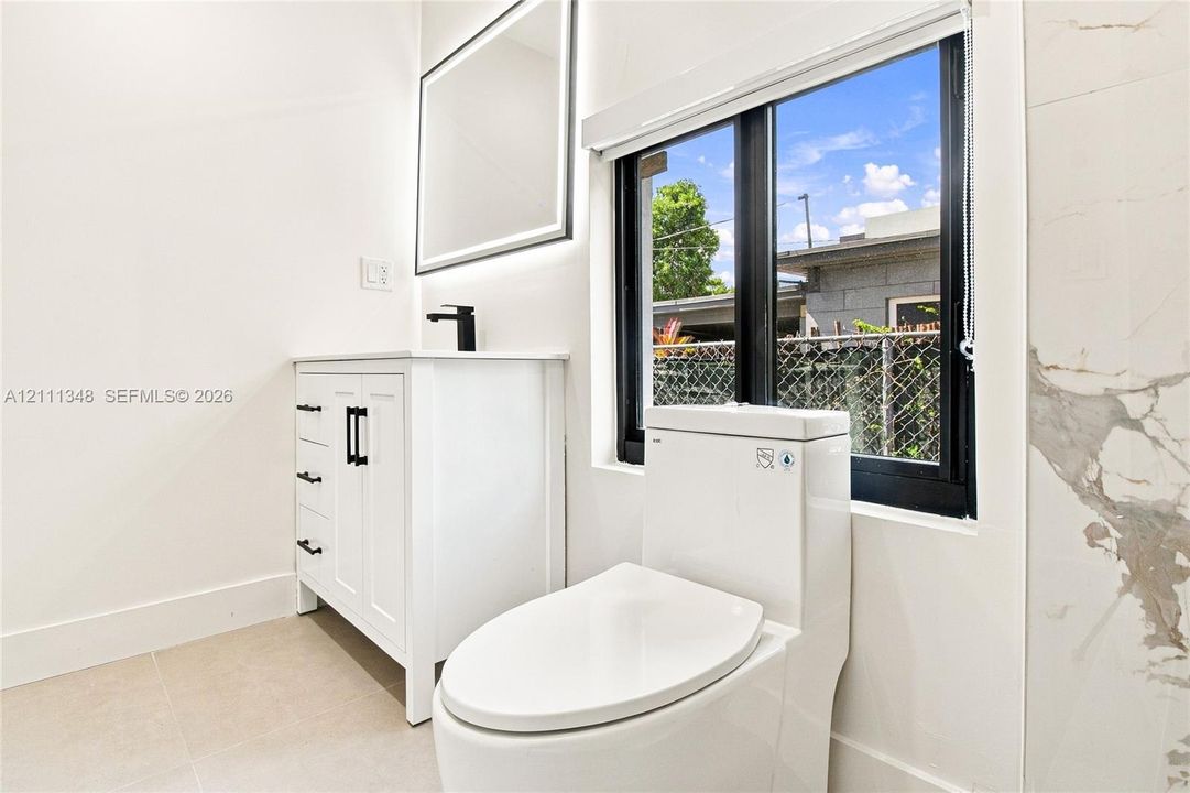 White vanity featuring black hardware, rectangular mirror with integrated lighting, matte black faucet, and porcelain tile wall surround