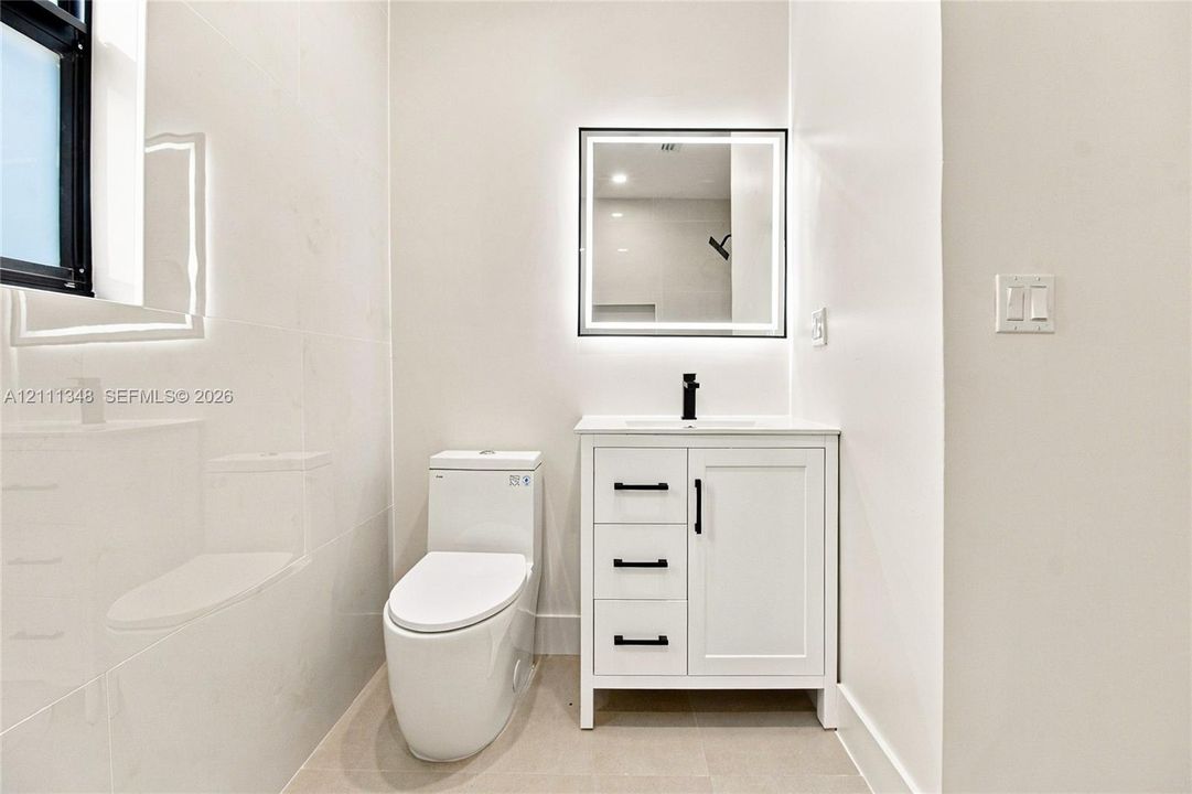 White cabinetry vanity with black hardware, integrated countertop with a matte black faucet, illuminated square mirror, one-piece toilet, and polished wall tile