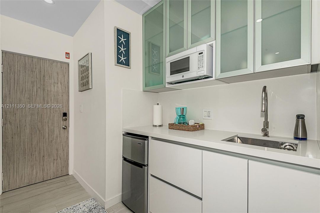 Kitchenette featuring white cabinetry with glass-panel upper doors, white solid-surface countertops, an integrated stainless steel sink with a gooseneck faucet, a built-in microwave, and wood-finish entry door