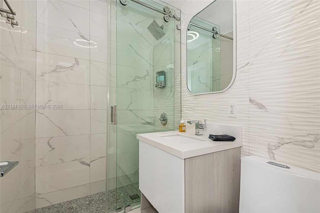 Walk-in shower featuring a sliding glass door, rainfall showerhead, and stone-tiled floor, accented by a floating vanity with a single-basin sink and a textured tile wall