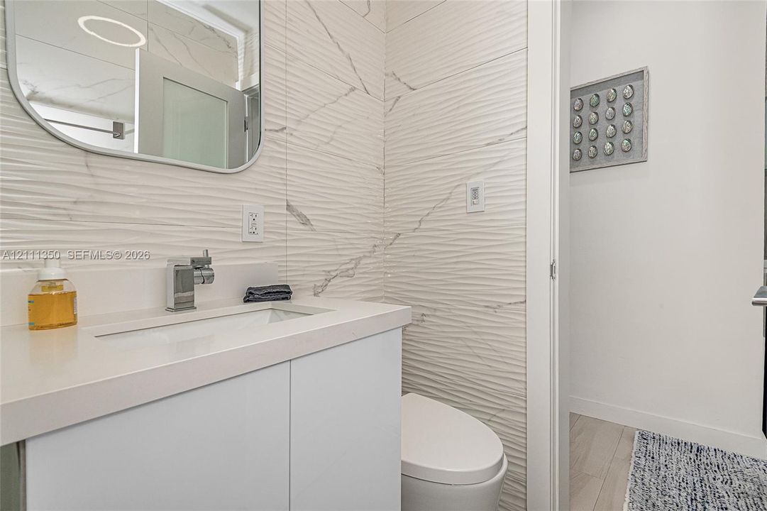 Modern vanity with stone-composite countertop and integrated sink, polished chrome faucet, rounded-edge wall mirror, and textured wall tile