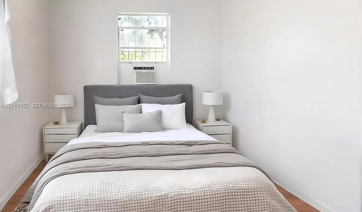 Neutral-toned interior space featuring wood-finish flooring, white wall surfaces, and a central window with an integrated wall-mounted cooling unit