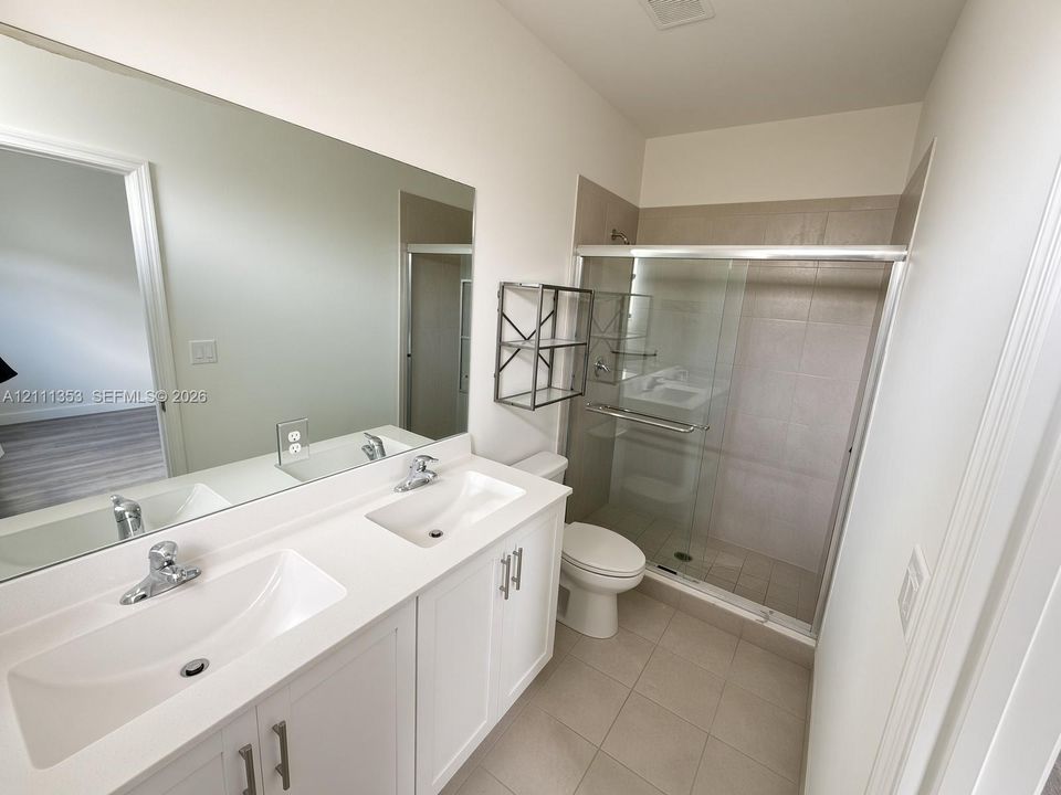 Dual-sink white vanity with solid surface countertops, frameless plate glass mirror, walk-in shower with glass enclosure, and ceramic tile flooring