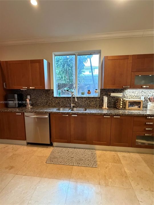 Kitchen featuring warm-toned wood cabinetry, natural stone countertops, a full-height stone backsplash, stainless steel dishwasher, and large-format tile flooring