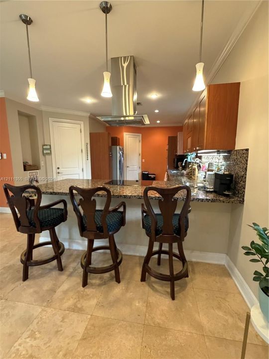 Kitchen featuring an expansive stone countertop, stainless steel ceiling-mounted vent hood, natural stone tile flooring, three pendant lights, and wood-finish cabinetry