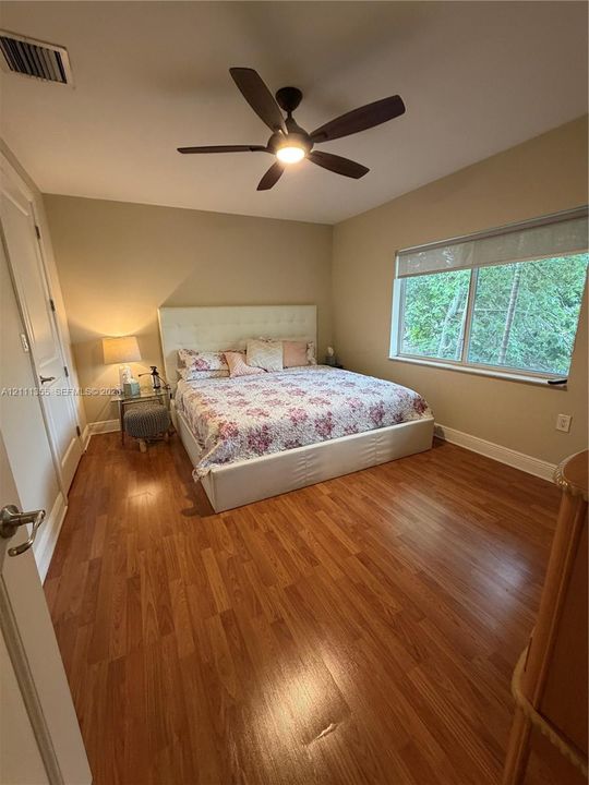 Room featuring wood-finish flooring, a multi-blade ceiling fan with integrated lighting, and a large window with a roller shade