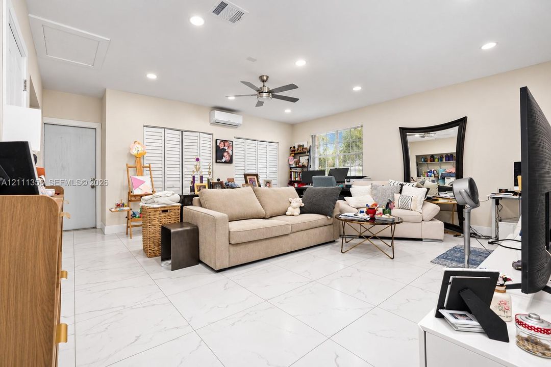 Living area featuring large-format tile flooring, recessed lighting, a ceiling fan, white louvered window shutters, and a wall-mounted ductless climate control unit