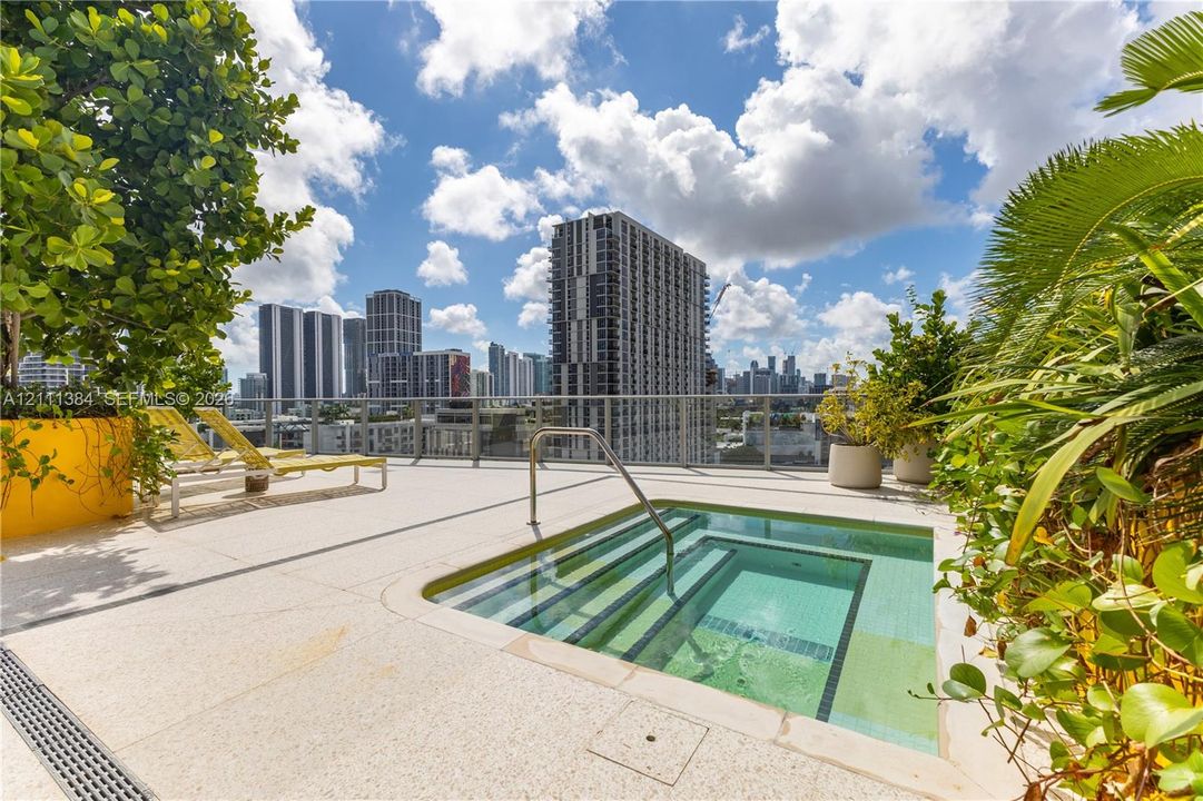 Rooftop terrace featuring an in-ground spa with integrated steps, a metal handrail, light-colored aggregate decking, and a glass-paneled perimeter railing