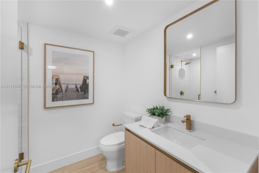 Modern vanity featuring a light wood-finish cabinet, white solid-surface countertop, and gold-toned faucet, paired with a rectangular gold-trimmed mirror and wood-finish flooring