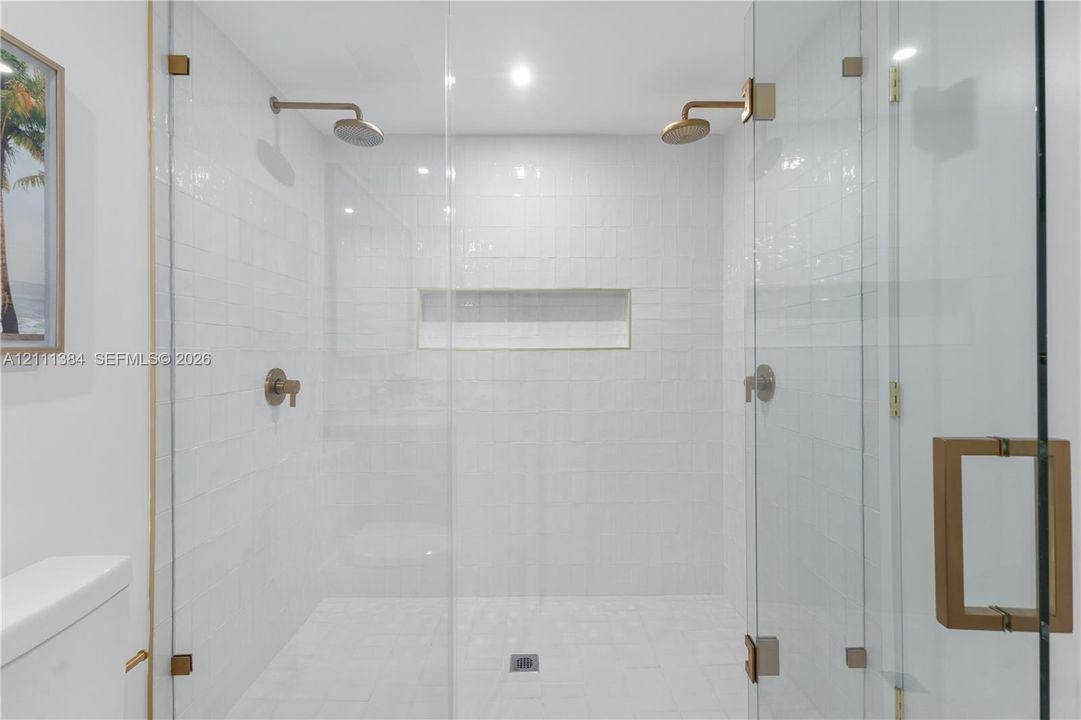 Walk-in shower featuring white tiled walls, dual rain-style showerheads, an integrated rectangular wall niche, and brass-finish hardware enclosed by a frameless glass door