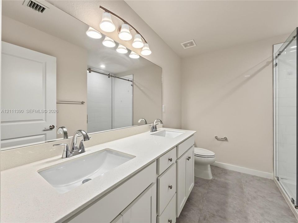 Dual-sink vanity with white cabinetry, light-toned stone countertops, polished chrome fixtures, full-width mirror, and a glass-enclosed shower