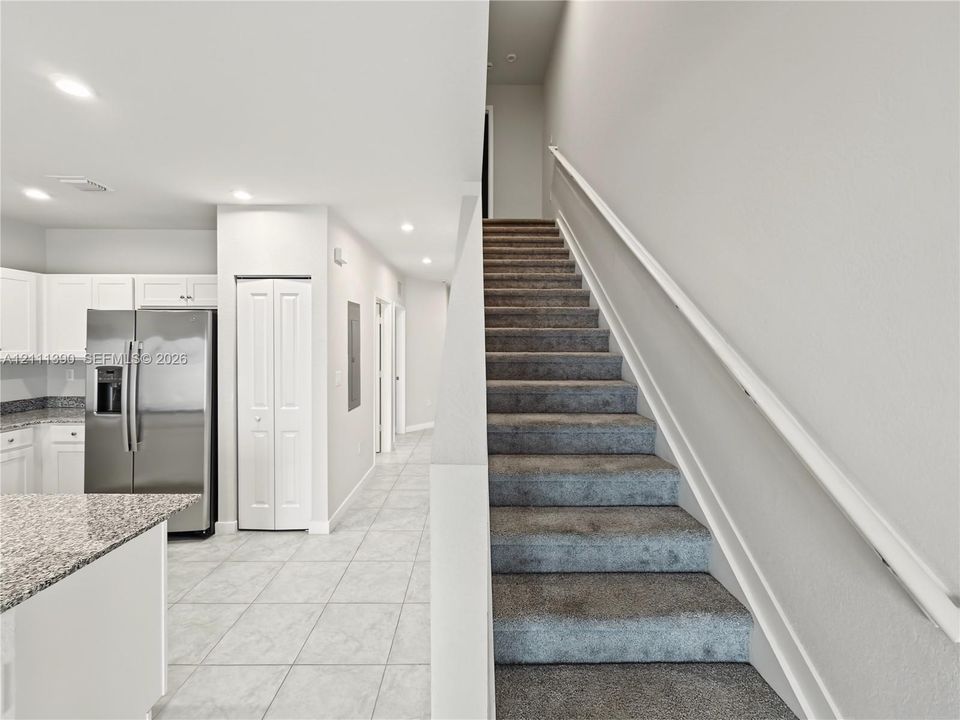 Kitchen featuring stainless steel refrigerator, white cabinetry, and stone-finish countertops, with tiled flooring and carpeted staircase with white handrail