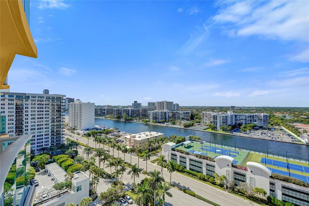 Elevated balcony perspective overlooking a waterway, paved streets, palm-lined grounds, and recreational courts