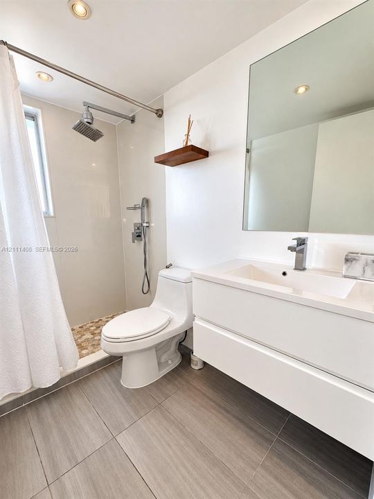 Floating white vanity with an integrated basin, wall-mounted rectangular mirror, wood-finish flooring, shower with a rainfall showerhead and handheld attachment, and built-in wood shelving