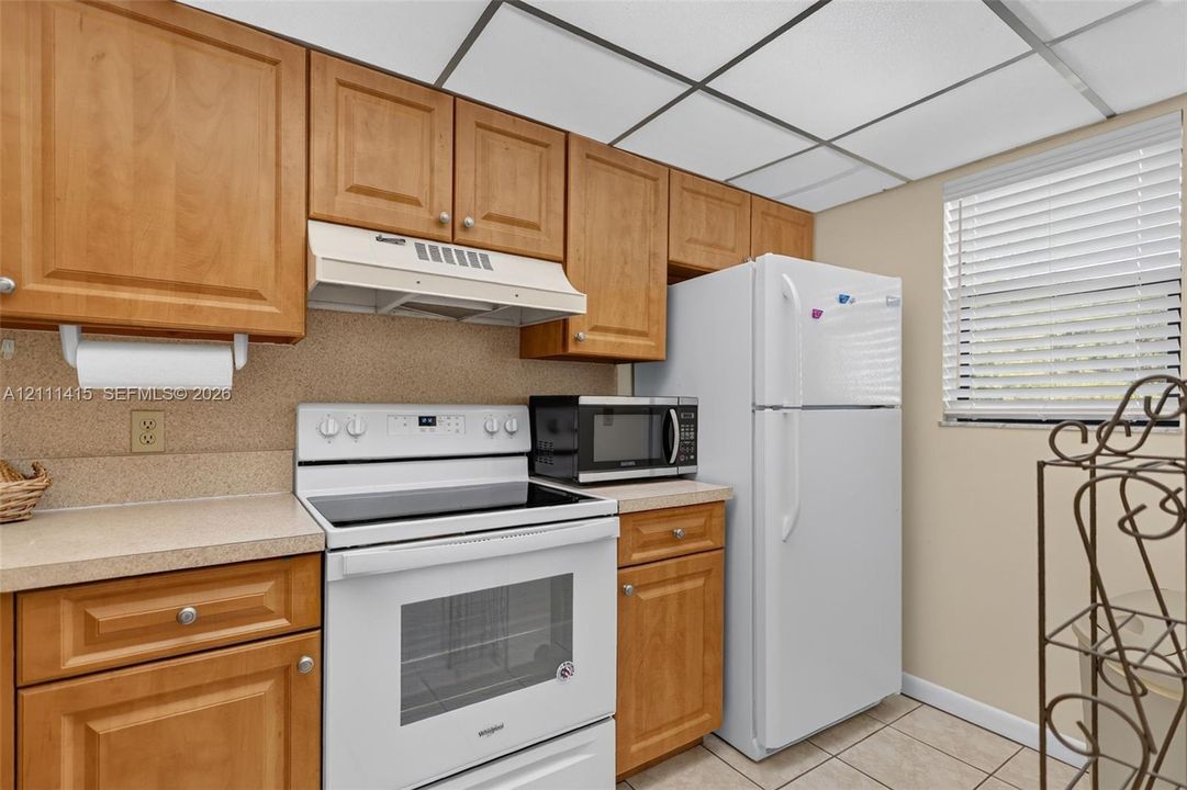 Kitchen featuring medium-tone wood cabinetry, solid-surface countertops, white electric range with built-in range hood, white refrigerator, and tile flooring