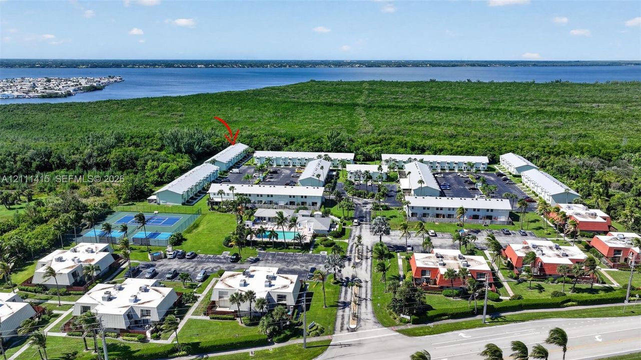 Aerial perspective of a multi-building residential complex featuring paved parking areas, a swimming pool, and a tennis court