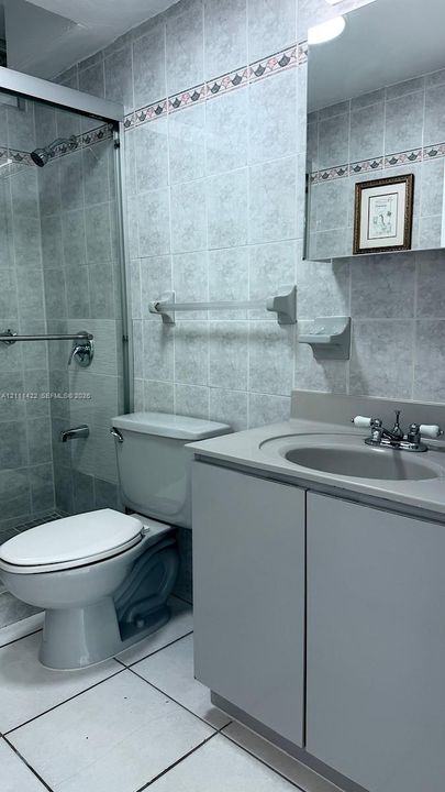 Bathroom featuring a grey vanity with an integrated basin, glass-enclosed shower, wall-mounted soap dish, metal towel bar, and floor-to-ceiling tilework with a decorative border
