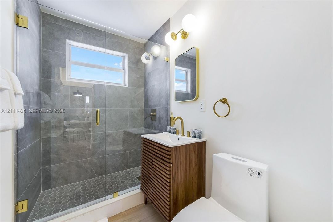 Walk-in shower featuring gray tile surround, glass enclosure, and hexagonal floor tile, complemented by a slatted wood-finish vanity, brass-toned fixtures, and a wall-mounted globe vanity light