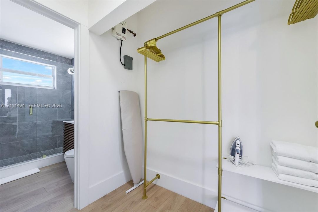 Storage area featuring gold-tone metal garment rods, white shelving, and wood-finish flooring, with a partial look at a bathroom containing large-format gray wall tile and a glass-enclosed shower