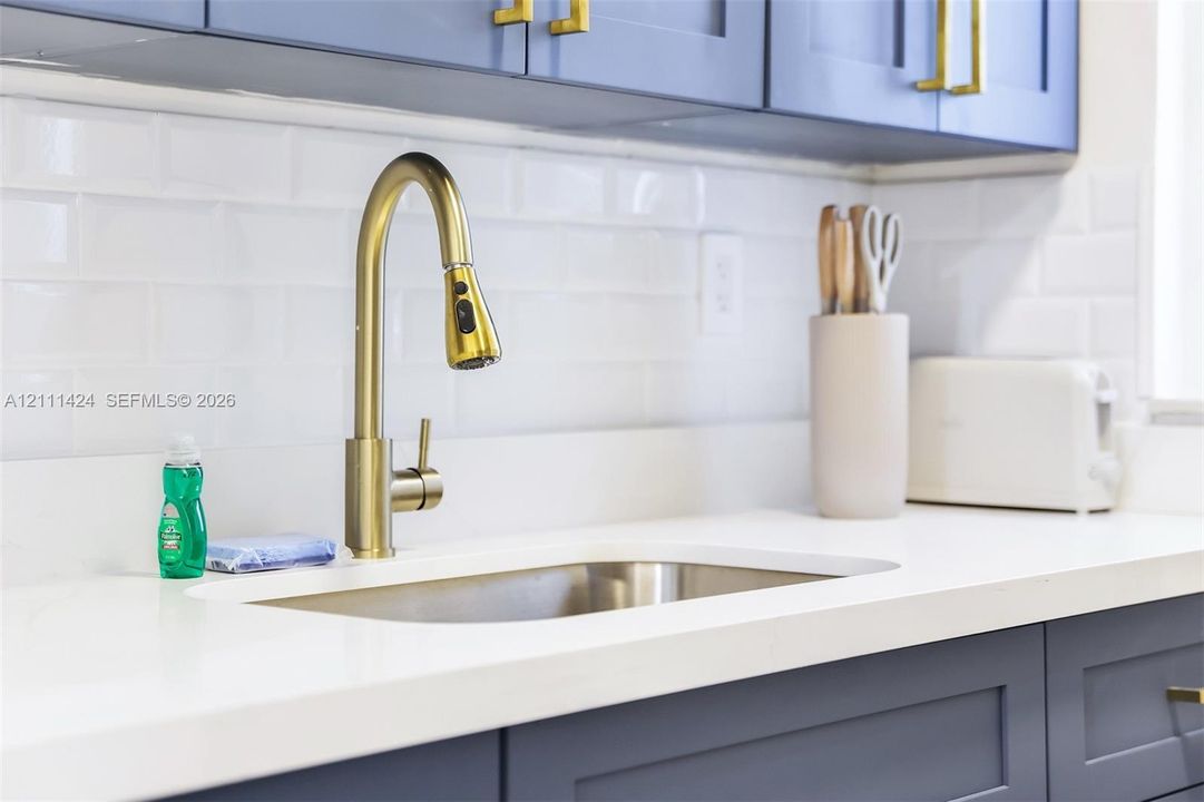 Blue shaker-style cabinetry with brass-finish hardware, undermount stainless steel sink, gold-tone gooseneck pull-down faucet, white solid-surface countertop, and white subway tile backsplash