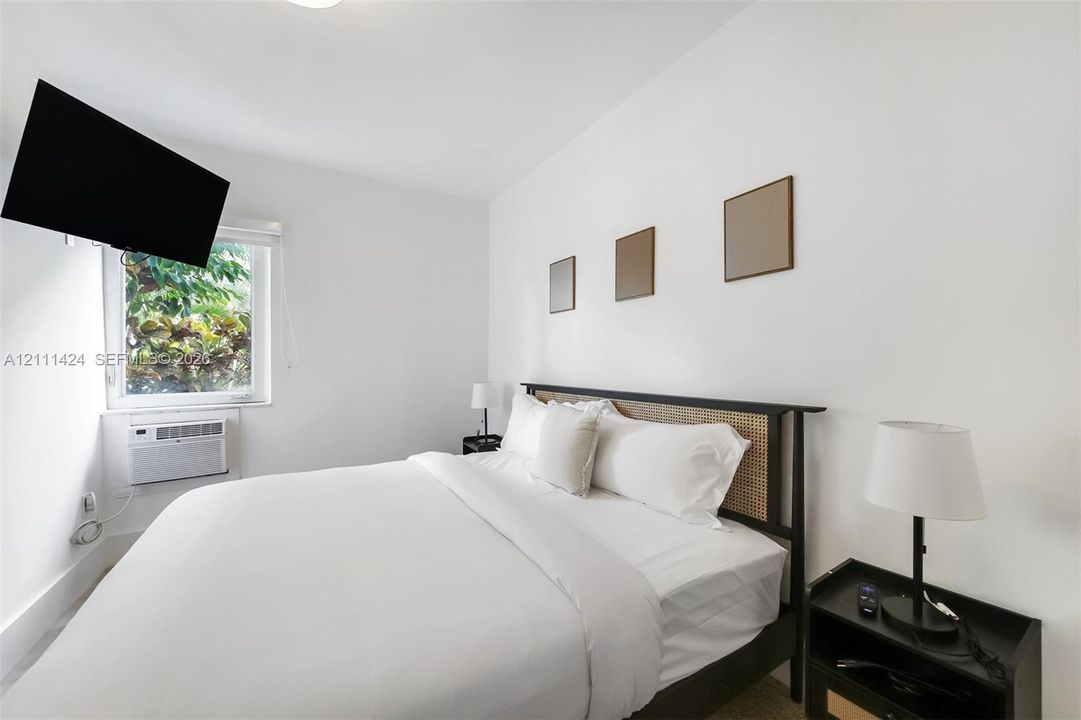 Bright bedroom featuring neutral walls, a single-pane window with exterior greenery, and an installed wall-mounted air conditioning unit