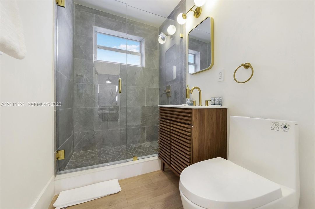 Walk-in shower with floor-to-ceiling stone-finish tile, glass enclosure, and wood-finish vanity featuring brushed brass fixtures, integrated wall-mounted lighting, and wood-finish flooring