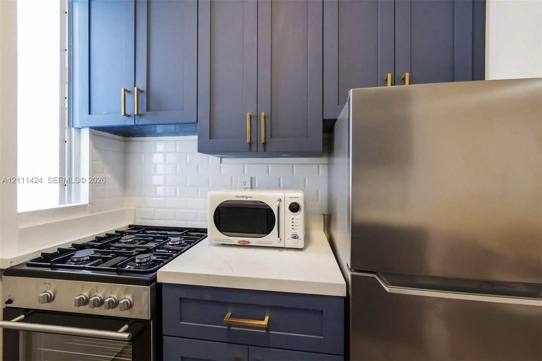 Kitchen featuring blue shaker-style cabinetry with brass-finish hardware, solid-surface countertops, white subway tile backsplash, and stainless steel appliances
