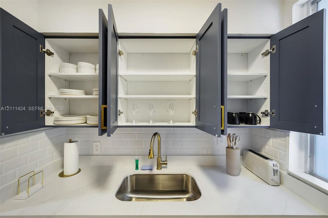 Shaker-style cabinetry with matte finish, white subway tile backsplash, solid-surface countertop, stainless steel undermount sink, and brass-finish gooseneck faucet