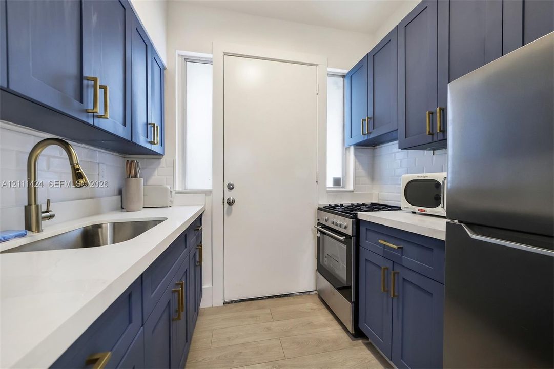 Navy blue cabinetry with brass-finish hardware, white composite countertops, white subway tile backsplash, stainless steel sink with pull-down faucet, and wood-finish flooring
