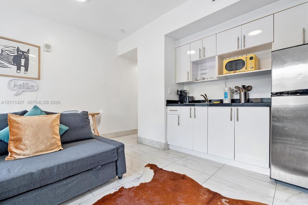 Living area with a sofa bed and large-format tile flooring, next to a kitchenette with white cabinetry, black countertops and a stainless steel refrigerator
