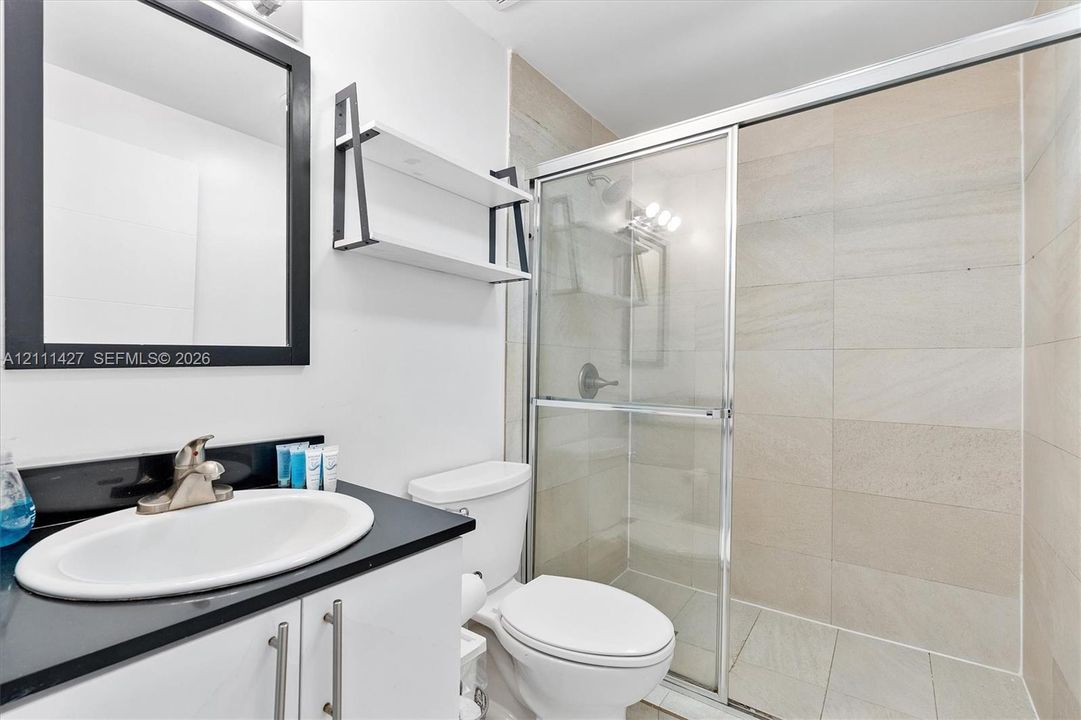 Bathroom featuring a vanity with a dark countertop and integrated sink, a framed mirror, a wall-mounted shelf, and a walk-in shower with a glass door and large-format wall tile