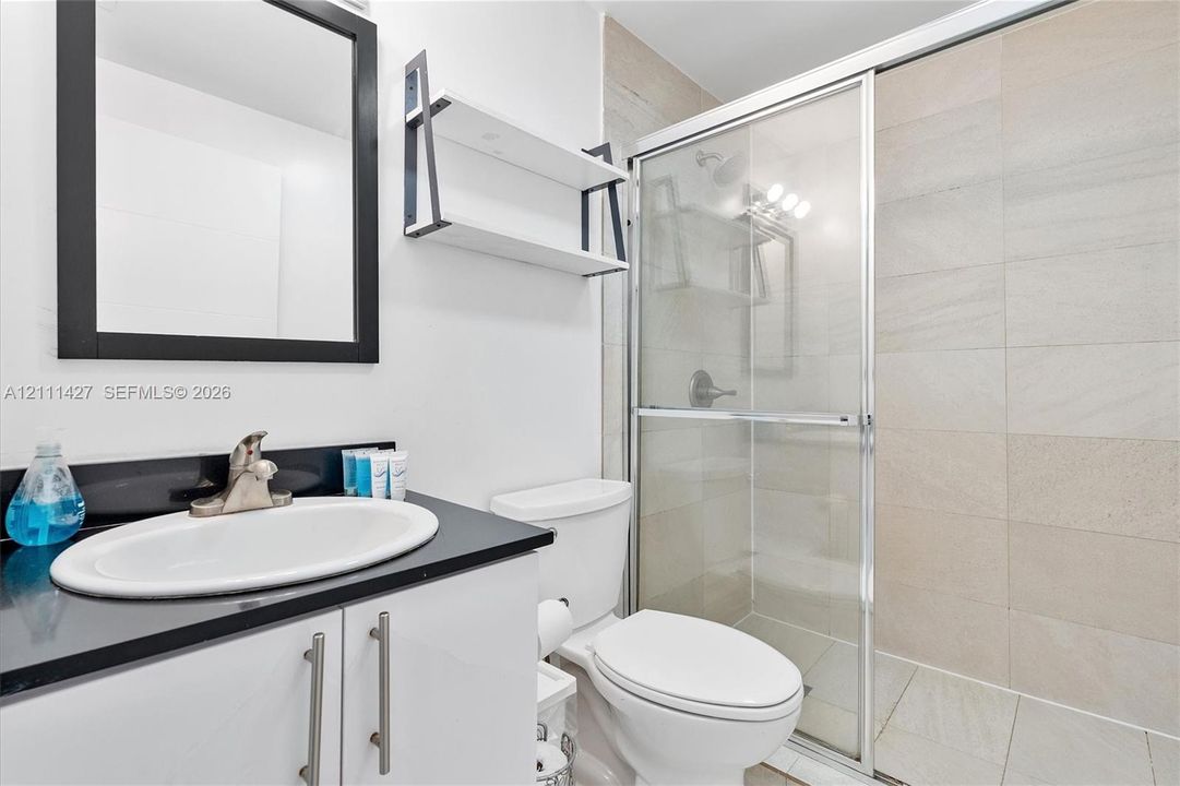 Bathroom featuring a vanity with a dark countertop and oval undermount sink, black-framed mirror, mounted shelving, and a shower enclosure with large-format wall tile and sliding glass door