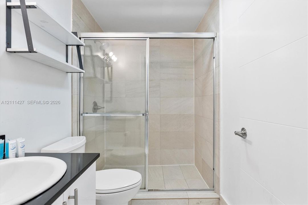 Walk-in shower featuring sliding glass doors, neutral tile surround, black countertop vanity, integrated shelving, and polished hardware