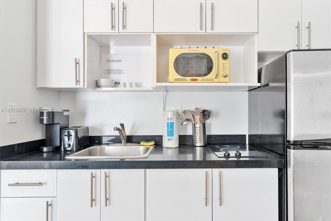 Kitchenette featuring white cabinetry with brushed-metal pulls, solid-surface dark countertops, a stainless steel undermount sink, and an integrated electric cooktop
