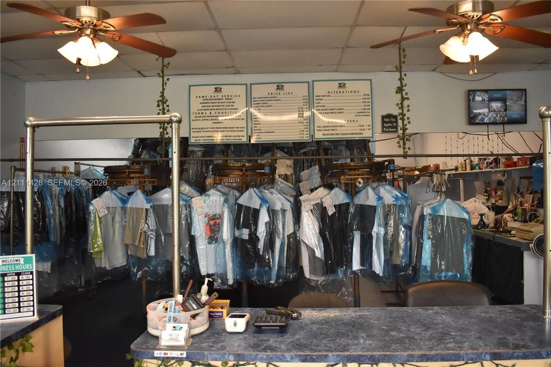Commercial interior featuring a service counter with a stone-finish top, ceiling-mounted paddle fans with integrated lighting, and dual overhead garment conveyor systems