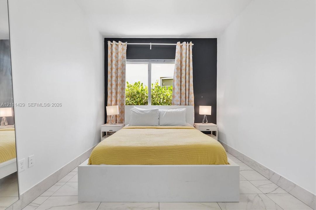 Bedroom interior featuring large-format tile flooring, a dark charcoal accent wall, a central window, and an integrated floor-to-ceiling mirror