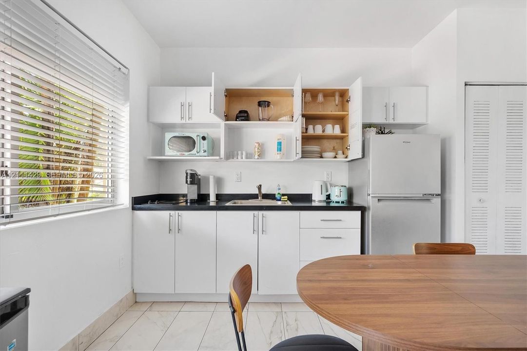 Kitchen featuring white shaker-style cabinetry, dark solid-surface countertops, stainless steel refrigerator, white tile flooring, and horizontal window blinds
