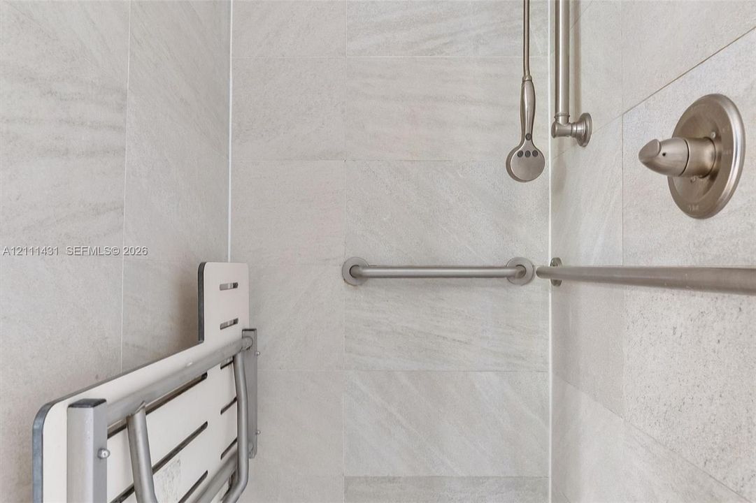 Shower stall featuring floor-to-ceiling light-toned stone tile, brushed nickel wall-mounted shower head, handheld sprayer, and integrated support bars