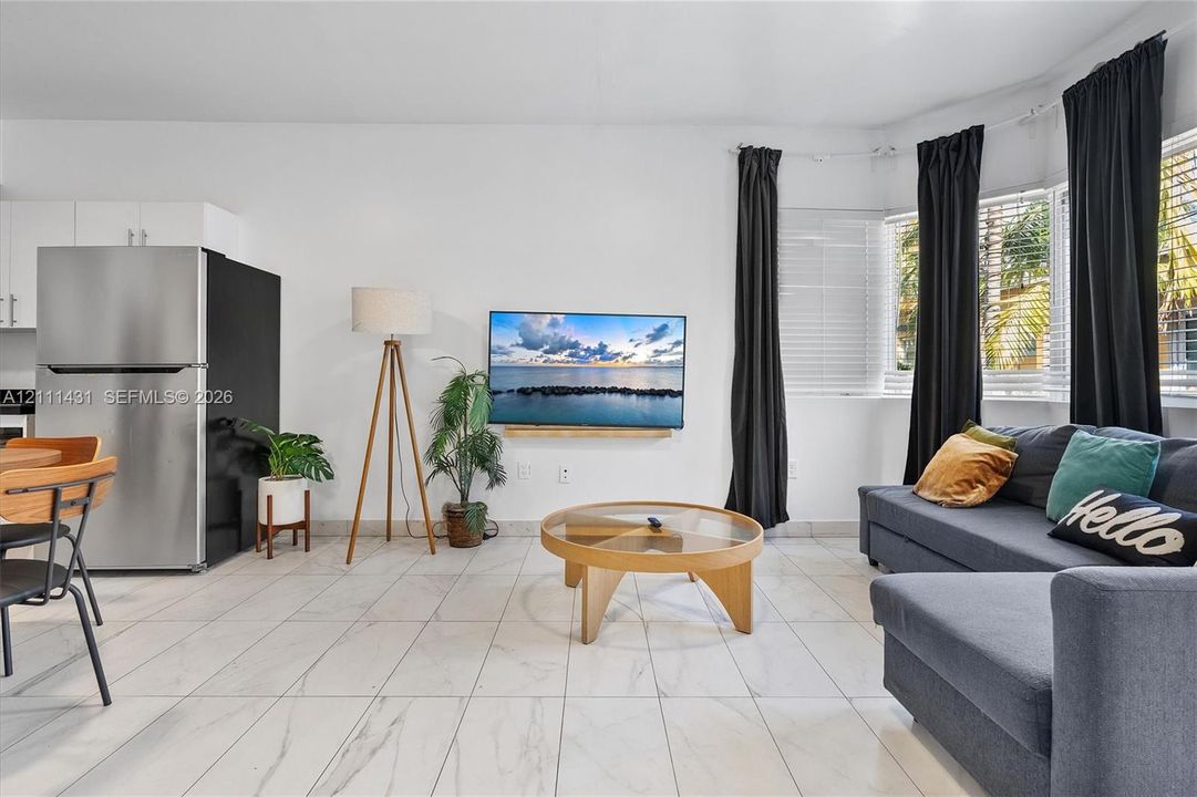 Living area featuring large-format tile flooring, a bay window with horizontal blinds, and a stainless steel refrigerator