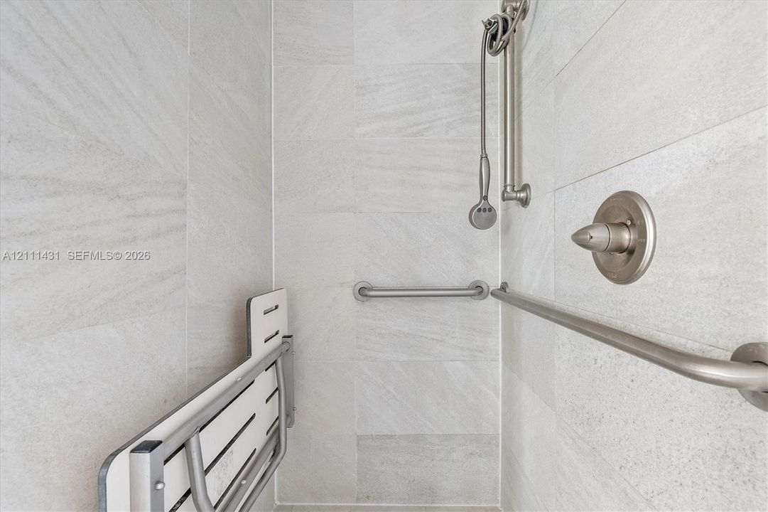 Shower enclosure featuring light grey stone-finish wall tiling, brushed nickel hardware, a wall-mounted shower head on a slide bar, and a fold-down wall-mounted seat