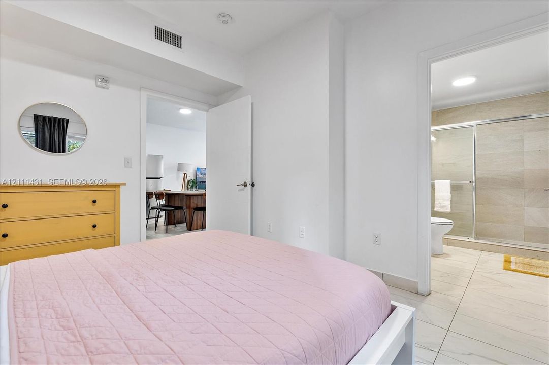 Bedroom with pink bedding, a yellow dresser and a round wall mirror, with a view through the doorway of a bathroom with a glass-enclosed shower