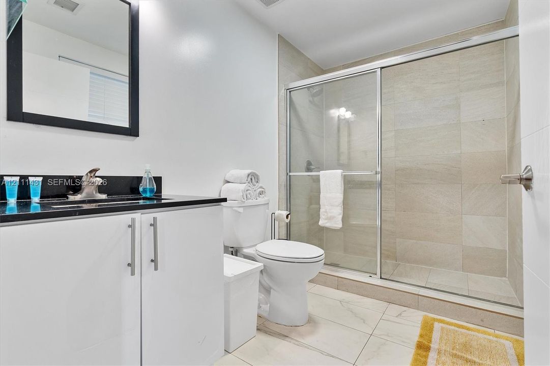 Bathroom featuring a vanity with dark stone countertop, rectangular undermount sink, walk-in shower with sliding glass doors, neutral tile wall surround, and large-format tile flooring