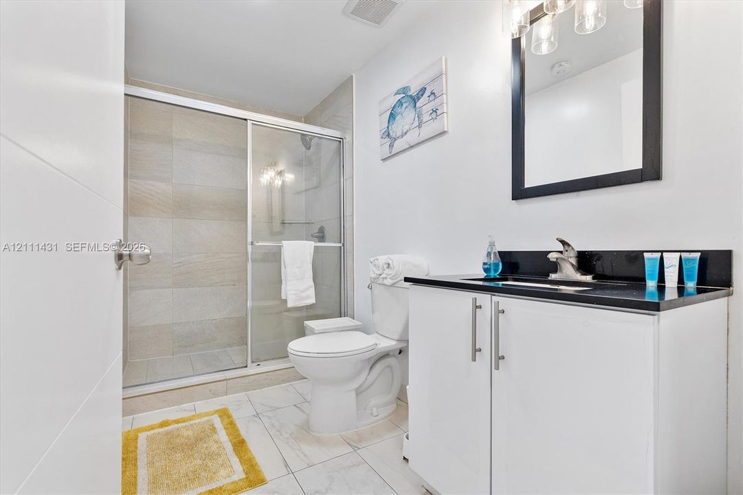 Bathroom featuring a white vanity cabinet with a dark stone countertop, an integrated sink, a framed mirror with integrated lighting, and a walk-in shower with a glass door and neutral wall tile