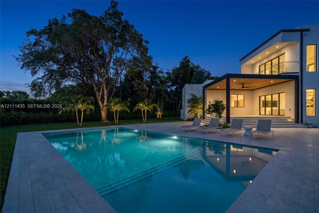 Modern architectural exterior featuring a swimming pool with integrated stairs, expansive stone paver pool deck, covered patio with wood-paneled ceiling, and second-story balcony with glass railing