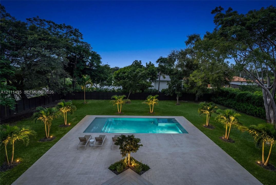 In-ground swimming pool featuring a tanning ledge, light-toned stone patio, manicured lawn, perimeter fencing, and established palms