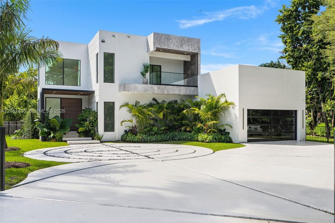 Modern architectural exterior featuring a smooth stucco facade, upper-level terrace with glass railing, decorative circular paver hardscaping, and a multi-panel glass garage door