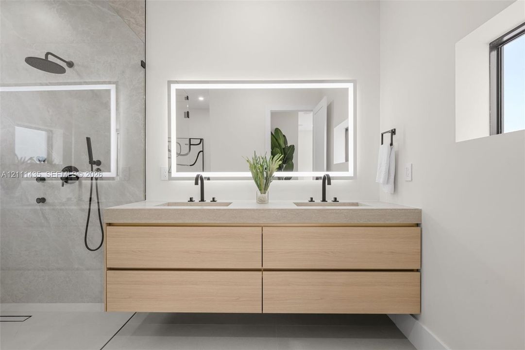 Floating wood-finish vanity with dual sinks, stone-surface countertop, LED-backlit mirror, and matte black fixtures, paired with a walk-in shower featuring large-format wall tile and matte black shower hardware